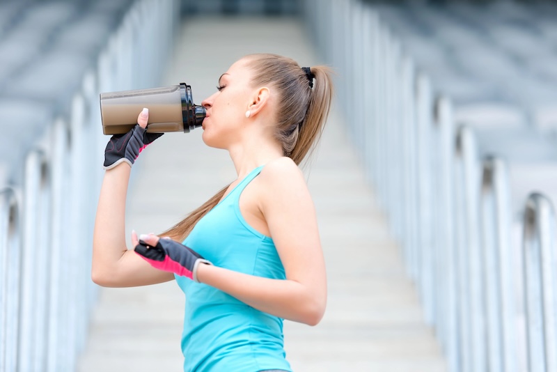 Active Woman Hydrating with Protein Shake Outdoors - Seaflora Skincare A woman in athletic gym wear sits on outdoor concrete steps, taking a break and drinking from a protein shake bottle on a sunny day.