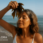 A woman in her 50s stands on a beach holding fresh seaweed in front of her face as golden seaweed oil gently drips onto her skin. Seaflora logo in white appears on the image.