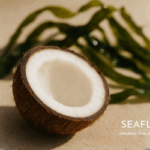 Alt Text A halved coconut sits on a sandy surface, with the interior visible. In the background, out of focus, is seaweed. The blog's logo is in the bottom right corner.